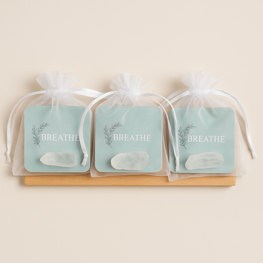 Three mini ritual pouches from Ara Candles are displayed in a row, each containing a square “Breathe” affirmation card in soft sage green with a sprig illustration, paired with a polished Amazonite crystal. The pouches are sheer white organza with satin drawstrings, resting on a wooden tray against a neutral beige background.