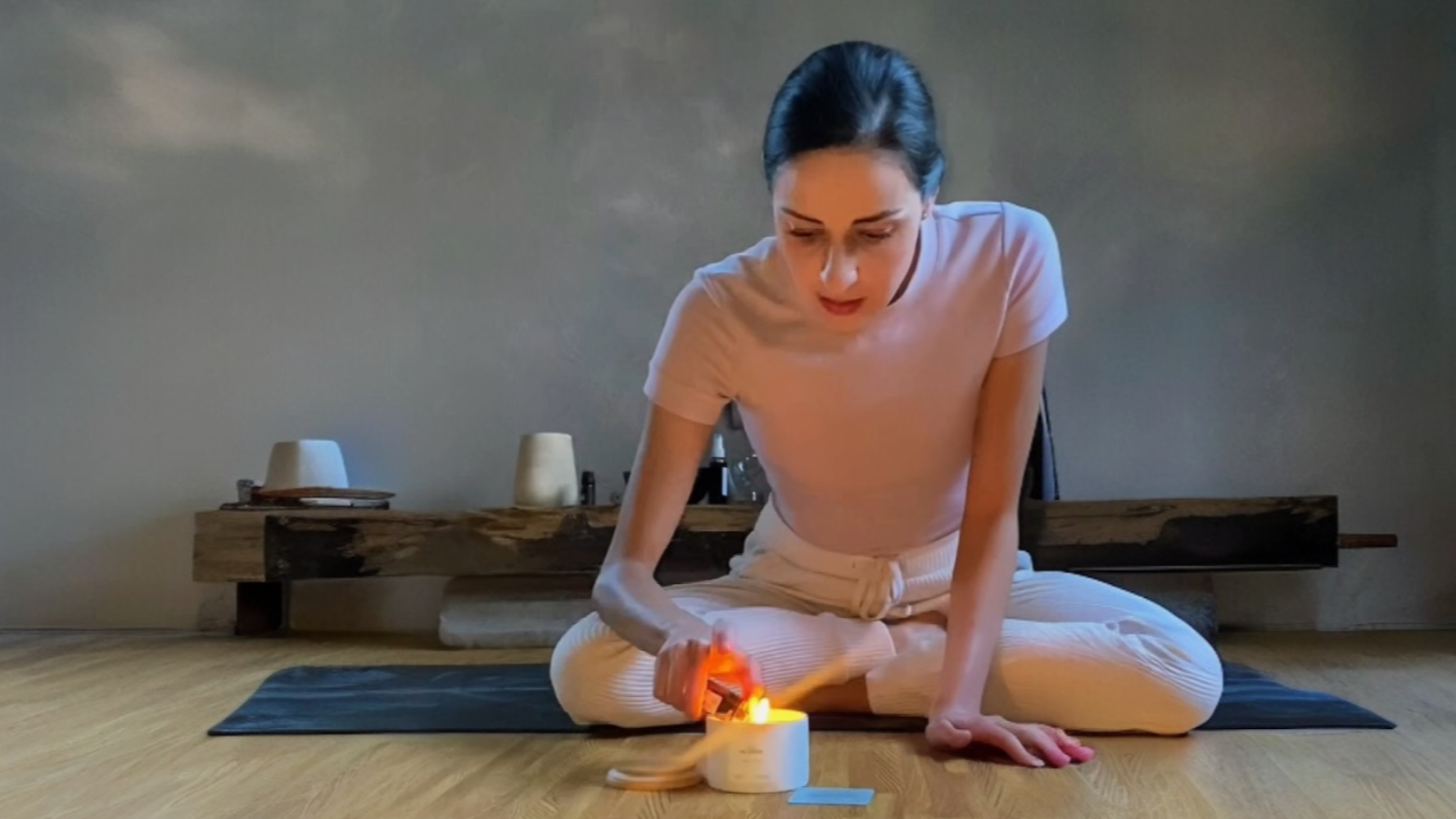 Woman in a white outfit sitting on a mat with candles and a bowl in a minimalistic room.