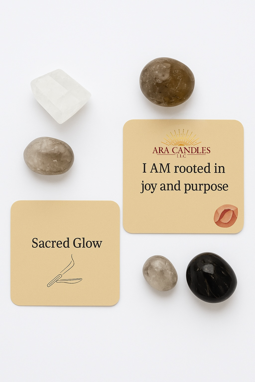 A styled photograph of two square affirmation cards from Ara Candles’ Sacred Glow scent. The top card reads “I AM rooted in joy and purpose” and features the Ara Candles LLC logo with a red-toned crystal illustration. The bottom card displays the scent name “Sacred Glow” with a minimal incense illustration. Crystals—Smoky Quartz, Clear Quartz, and two darker grounding stones—are artfully arranged around the cards on a clean white background.
