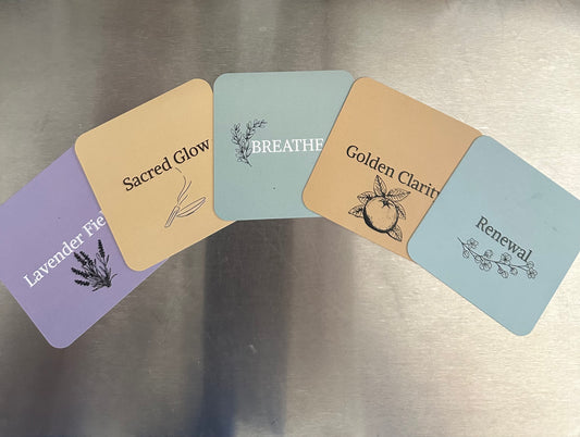 A fan arrangement of five Ara Candles affirmation cards displayed on a metallic surface, each representing a scent in the Wellness Collection. From left to right: Lavender Fields (lavender illustration, purple background), Sacred Glow (incense illustration, earthy tan), Breathe (sage sprig, soft green), Golden Clarity (citrus illustration, golden yellow), and Renewal (floral branch, pale green).