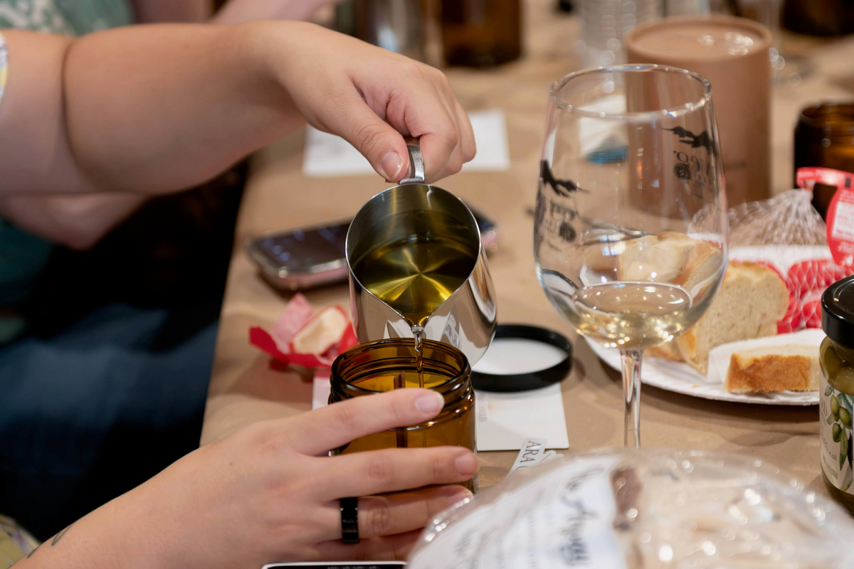 A close-up lifestyle shot of a person pouring golden olive oil from a metal pitcher into an amber glass jar on a rustic wooden table, surrounded by baking ingredients like butter, bread, and wine glasses.