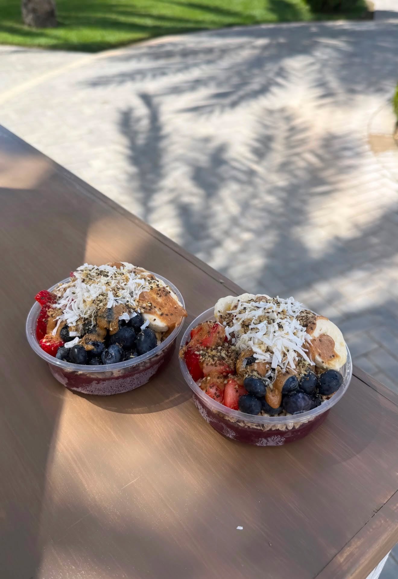 Two clear plastic bowls of fruit and nut smoothie or acai bowls topped with shredded coconut, placed on a wooden table outdoors with dappled sunlight and shadows.