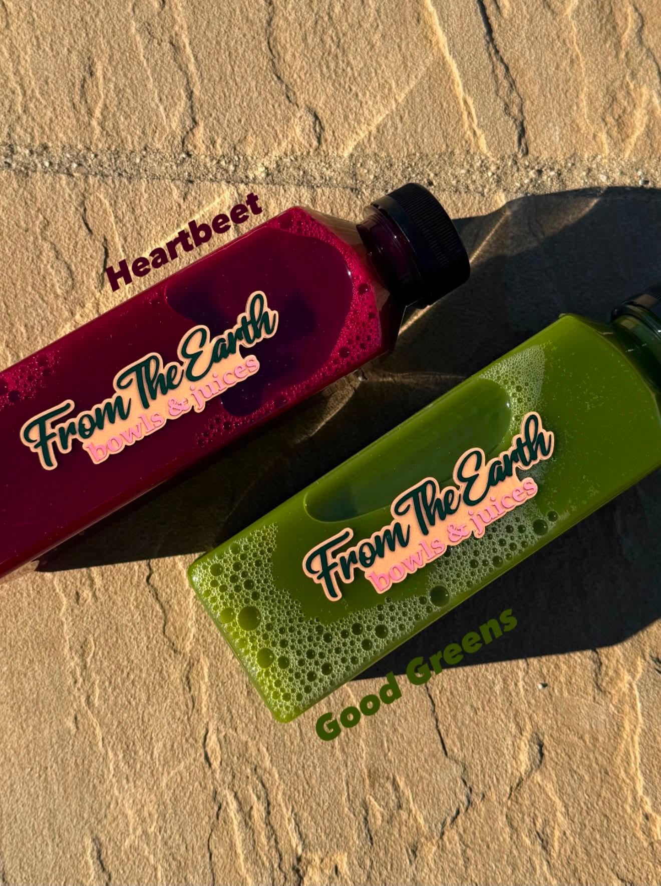 Two frosted plastic juice bottles from From The Earth Juice Co. labeled 'Heartbeat' (deep magenta/purple) and 'Good Greens' (lime green), placed diagonally on a textured sandy background.