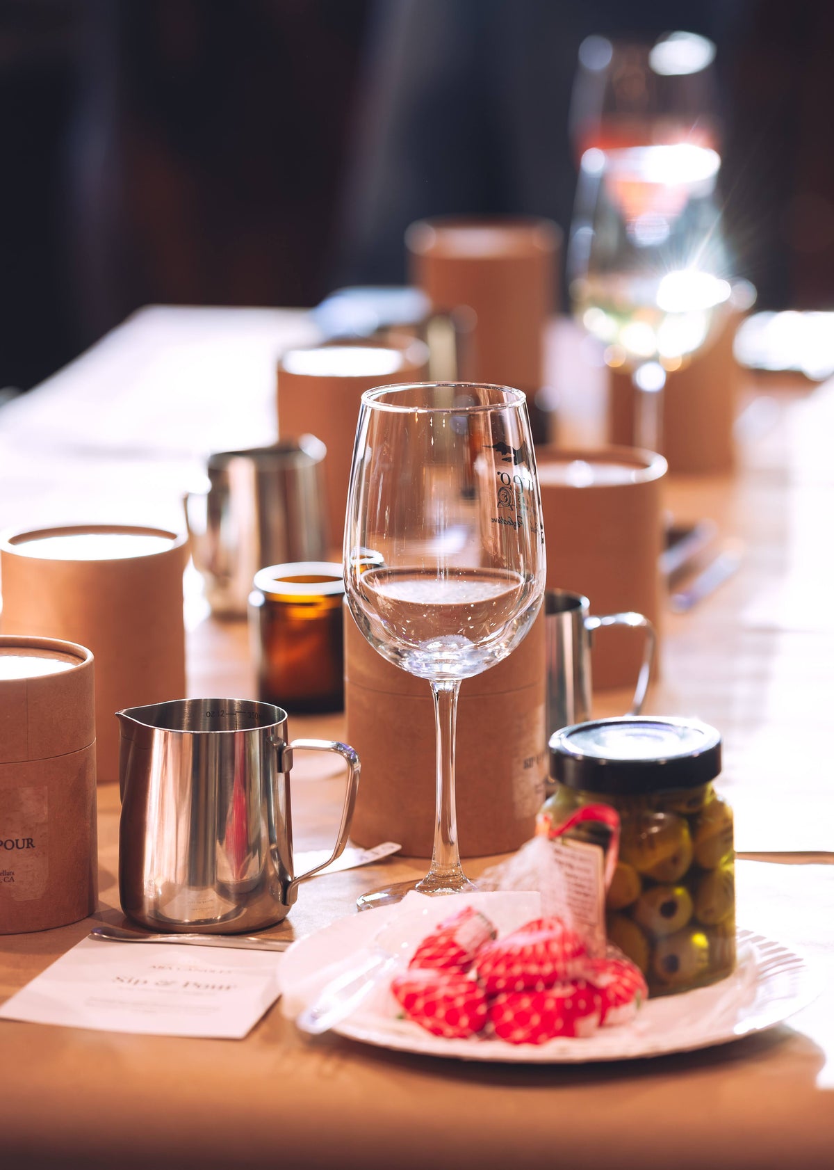 A styled dining table setting with a clear wine glass in the foreground, surrounded by brown paper candle sleeves, stainless steel mugs, and a small plate with red patterned napkins and a jar of olives. The background is softly blurred with warm lighting.