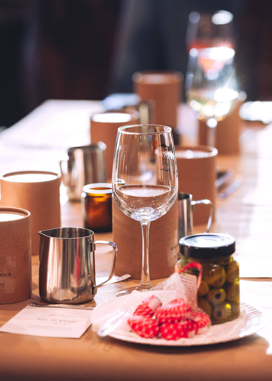 A styled dining table setting with a clear wine glass in the foreground, surrounded by brown paper candle sleeves, stainless steel mugs, and a small plate with red patterned napkins and a jar of olives. The background is softly blurred with warm lighting.