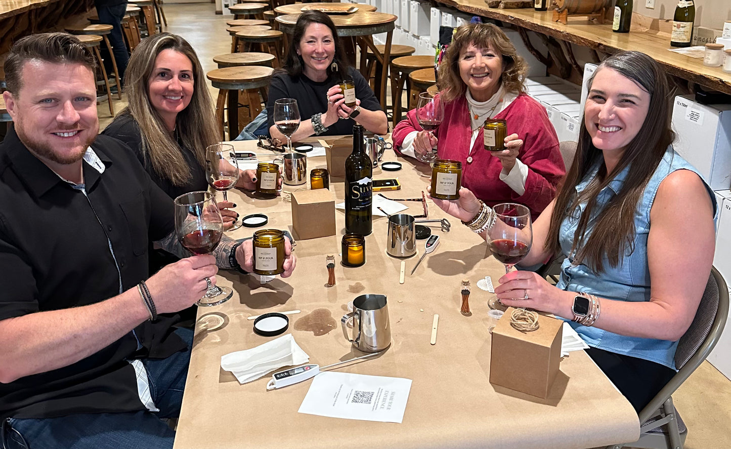 Group of people sitting around a table with wine glasses and bottles, possibly at a tasting event.