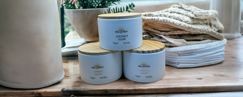 Three white ceramic summer candles from ARA candles- Coconut Glow, Citrus Agave, and Apricot Grove- stacked together on a wooden surface with soft natural light. 