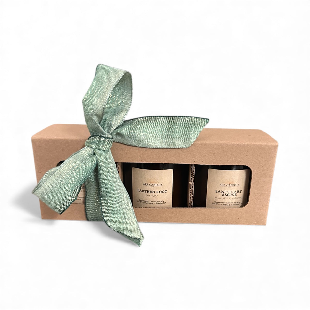 A serene Ara Candles Apothecary Collection gift set featuring two hand-poured wellness candles in amber jars, nestled in a kraft box tied with a soft green ribbon. Created as the Sacred Anchors trio, this set supports grounding, protection, and mindful ritual for conscious women seeking calm and sacred presence.