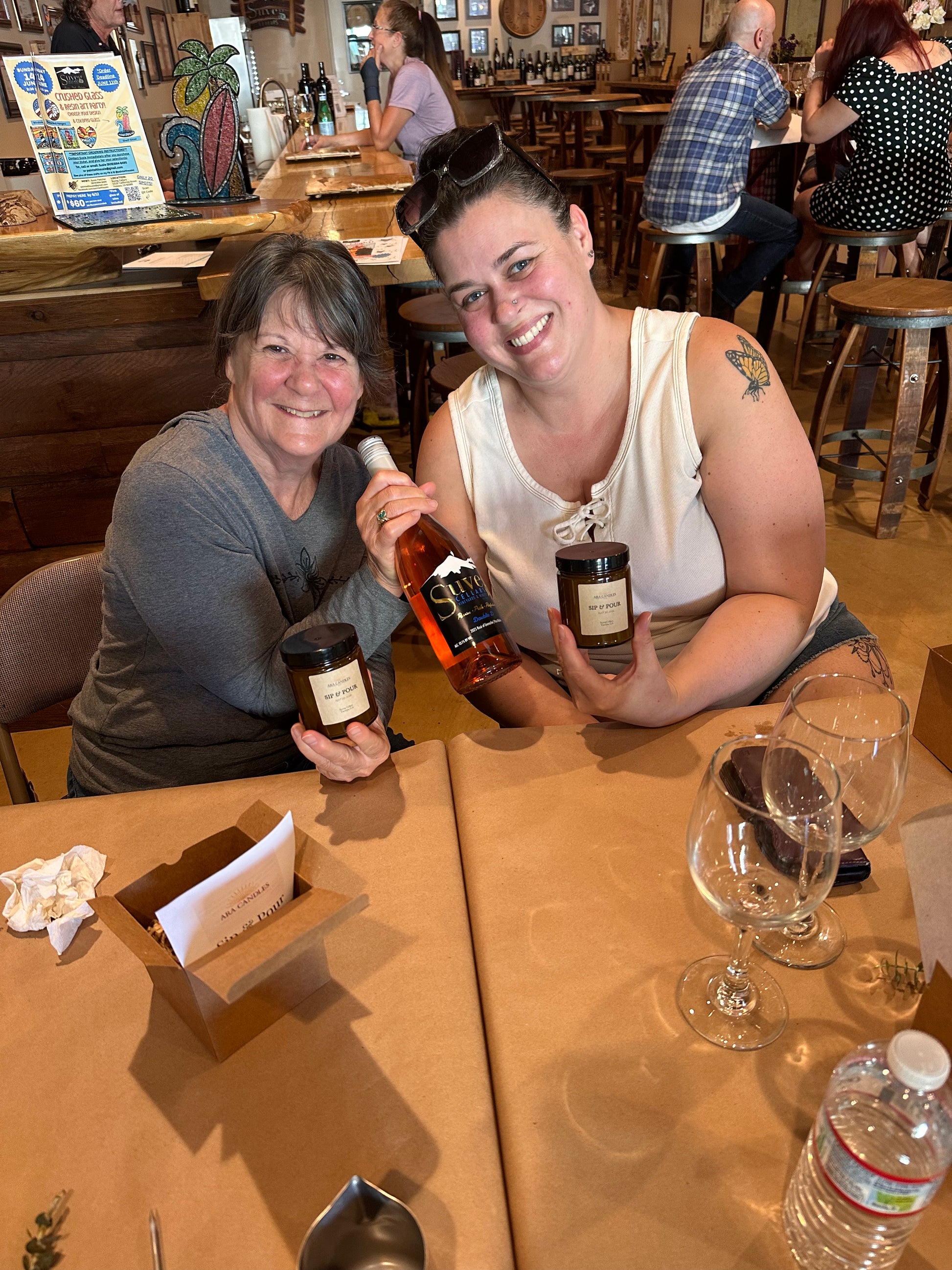 Two women sitting at a wooden table in a bar or restaurant setting, each holding a small jar and a bottle of what appears to be a beverage or sauce. The table has wine glasses, napkins, and a water bottle, with other people and bar fixtures visible in the background.