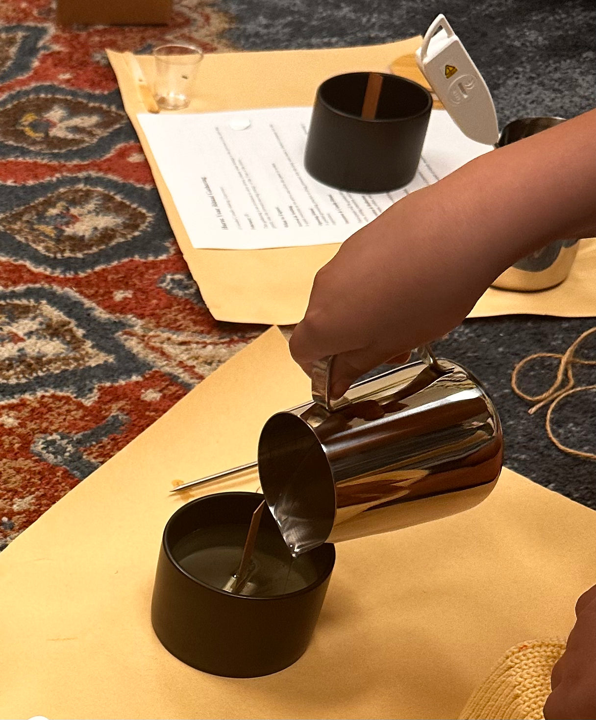 Person pouring wax from a silver pitcher into a black container on a table with a patterned rug.
