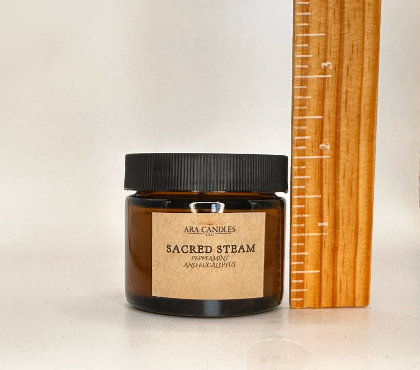 Candle jar labeled 'Sacred Steam' by Ara Candles next to a wooden ruler on a light background