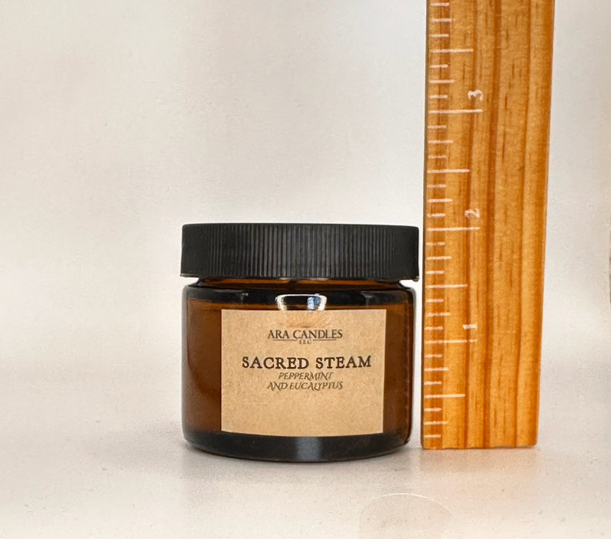 Candle jar labeled 'Sacred Steam' by Ara Candles next to a wooden ruler on a light background
